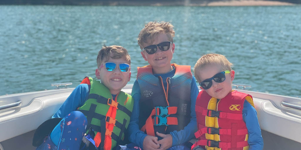 Three kids on a boat wearing Optic Nerve kids sunglasses and life vests with a lake in the background.