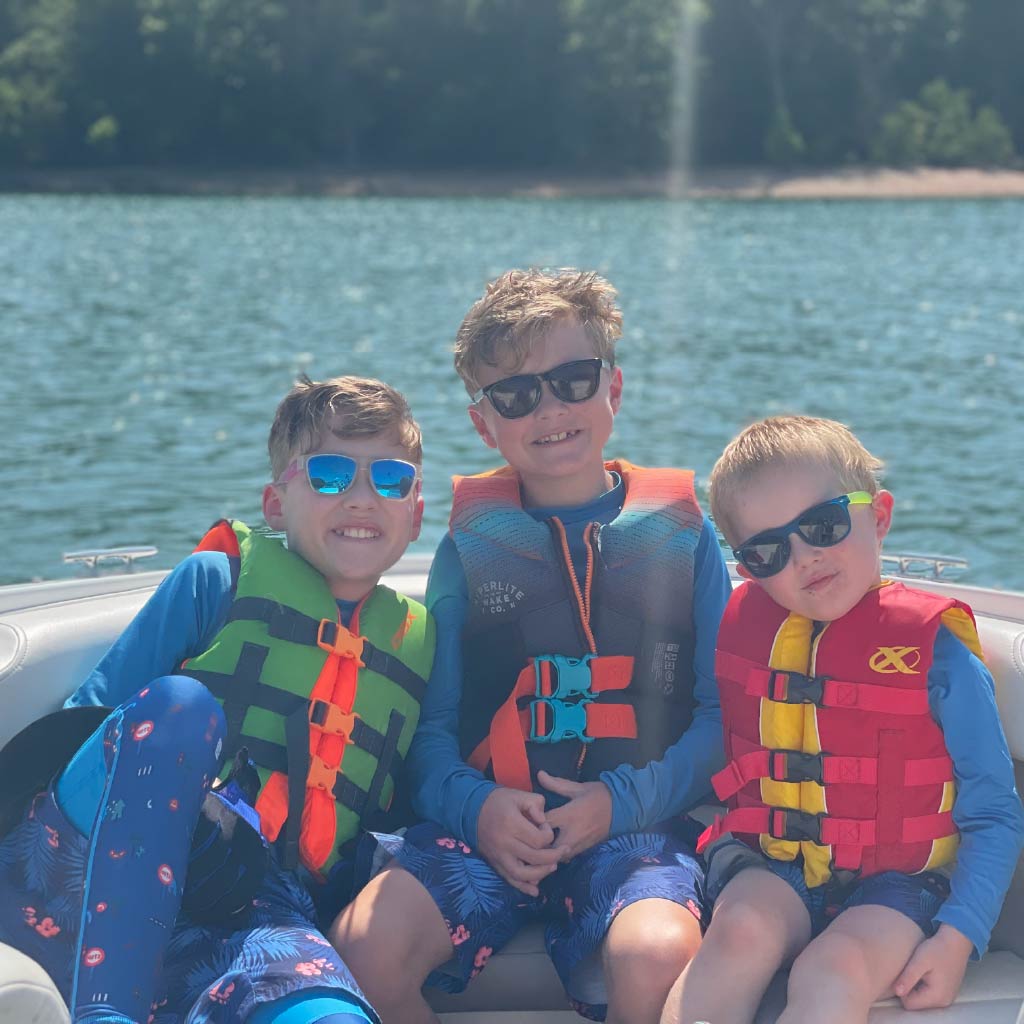 Three kids on a boat wearing Optic Nerve kids sunglasses and life vests with a lake in the background.