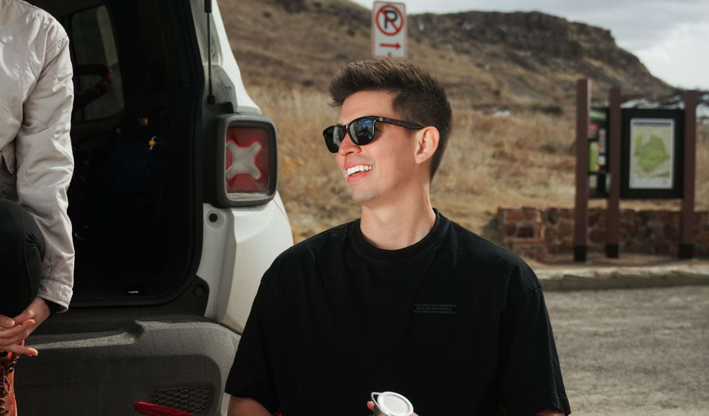 Man wearing a black shirt and Optic Nerve Fairplay sunglasses with a tortoise frame, smoke lenses, and a silver mirror, in front of the trunk of a car and an trailhead
