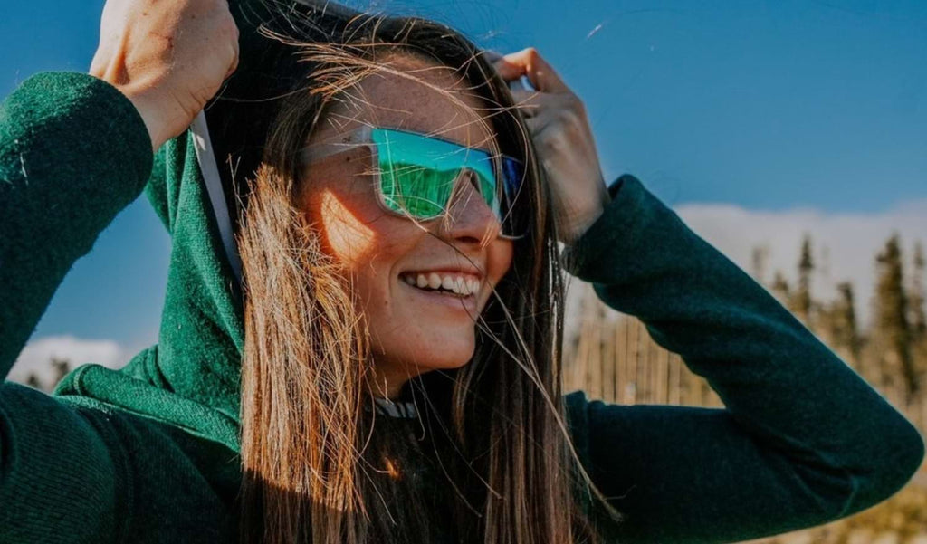 Woman wearing Optic Nerve Mojo Filter sunglasses with a clear frame, smoke lens, and a green mirror with blurred trees in the background