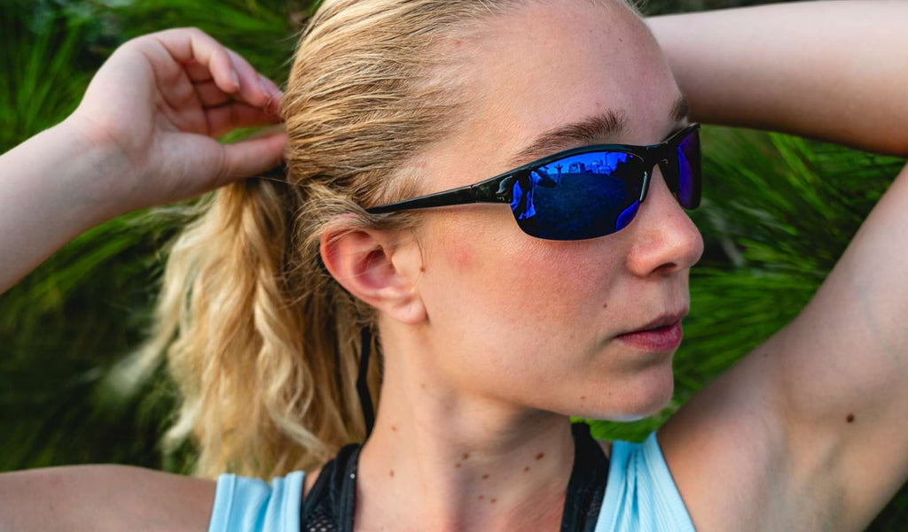 Woman wearing Optic Nerve Alpine sunglasses with black frame, smoke lenses, and blue mirror with a green blurred background