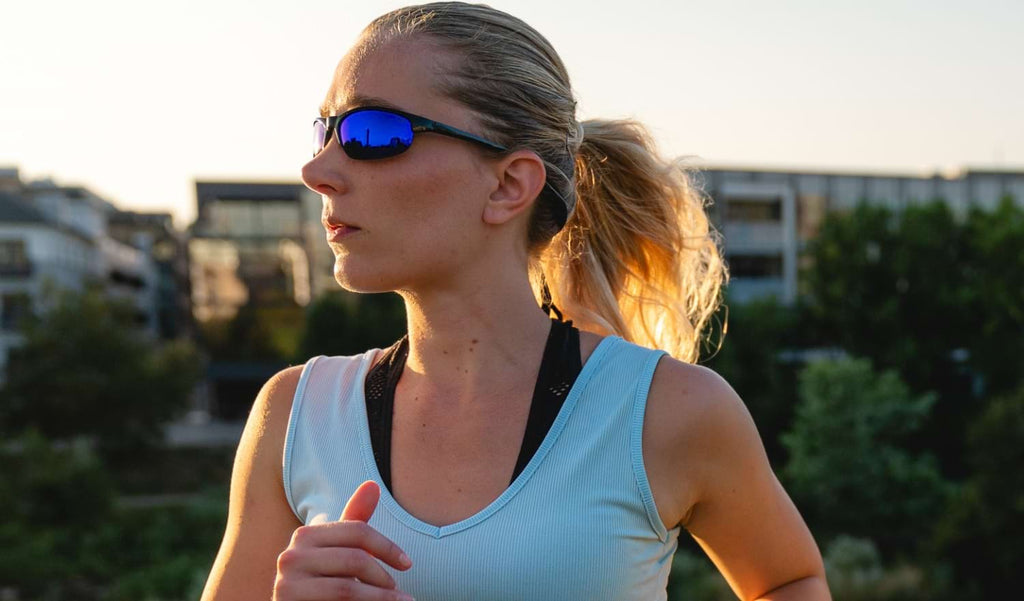Woman running wearing Optic Nerve Alpine sunglasses with a black frame, smoke lenses, and blue mirror with blurred buildings and trees in the background