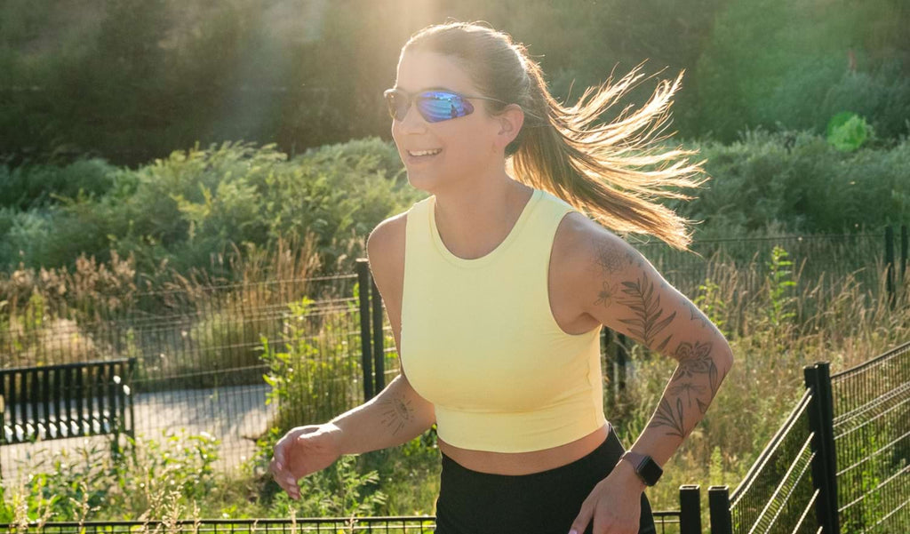 Woman running outside wearing Optic Nerve Alpine sunglasses with black frame, smoke lenses, and blue mirror with greenery background