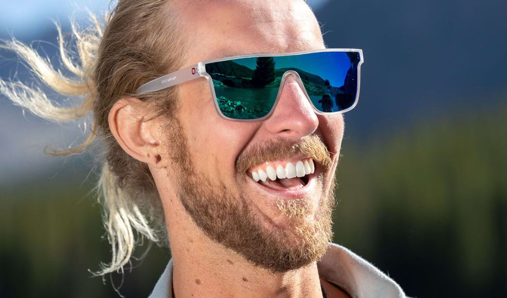 Close up of a man wearing Optic Nerve Mojo Filter sunglasses with a clear frame, smoke lens, and a green mirror with a blurred background
