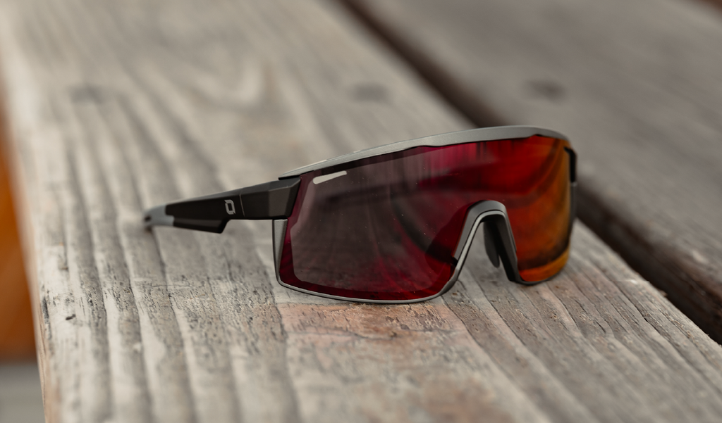 Optic Nerve FixieMAX sunglasses with a black frame, smoke lens, and red mirror on a wooden surface