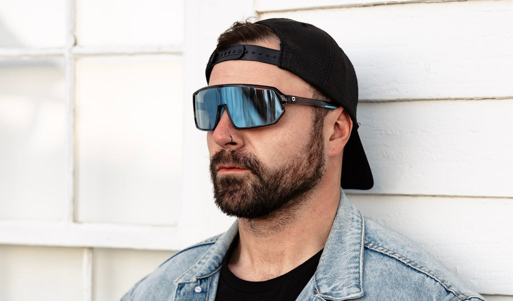 Man wearing a black baseball cap and Optic nerve FixieRIP sunglasses with a gray frame, smoke lens, and a blue mirror in front of a white wall
