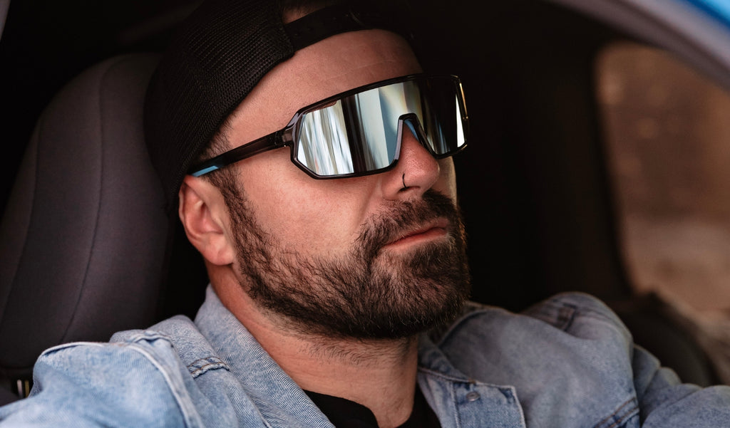 Man wearing Optic Nerve FixieRIP sunglasses with gray frame, smoke lens, and blue mirror inside a vehicle