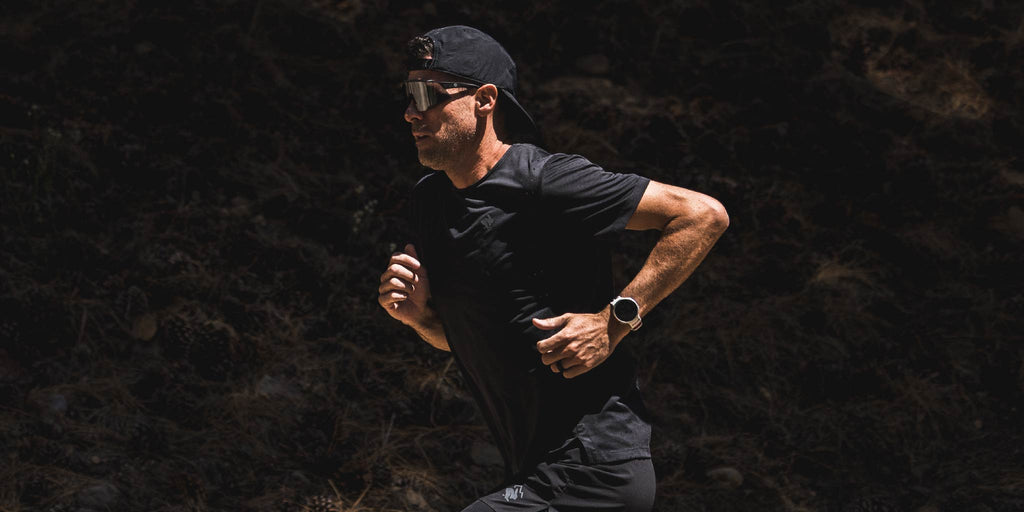Man running on a dark forest path wearing a black t-shirt, cap, and Optic Nerve FixieAIR sunglasses with a black frame, smoke lens, and a silver mirror.