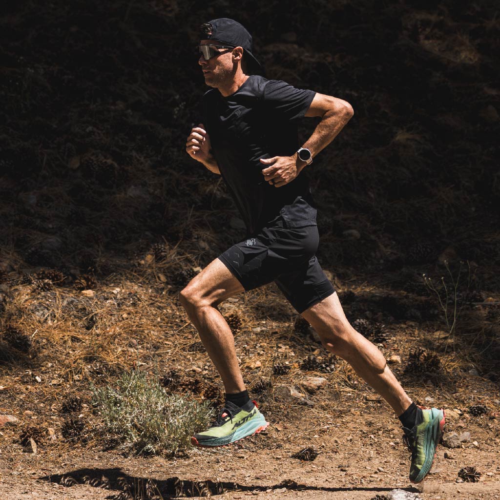 Man running on a trail in a forested area wearing all black and Optic Nerve FixieAIR sunglasses with a black frame, smoke lens, silver mirror.