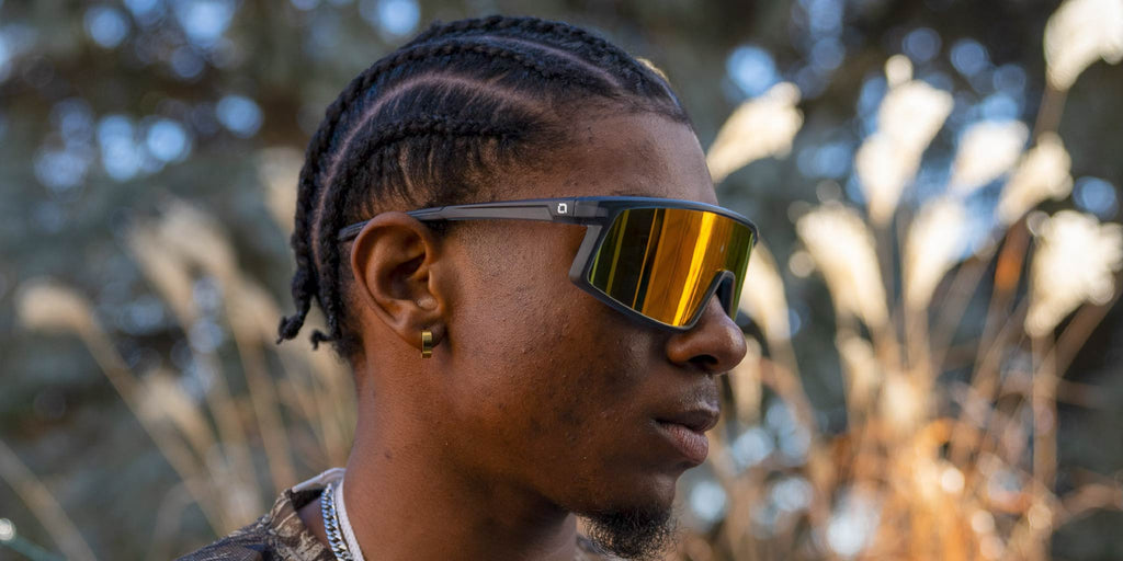 Person wearing Optic Nerve Stone Cat sunglasses with a gray frame, smoke lens, and red mirror, with a blurred natural background