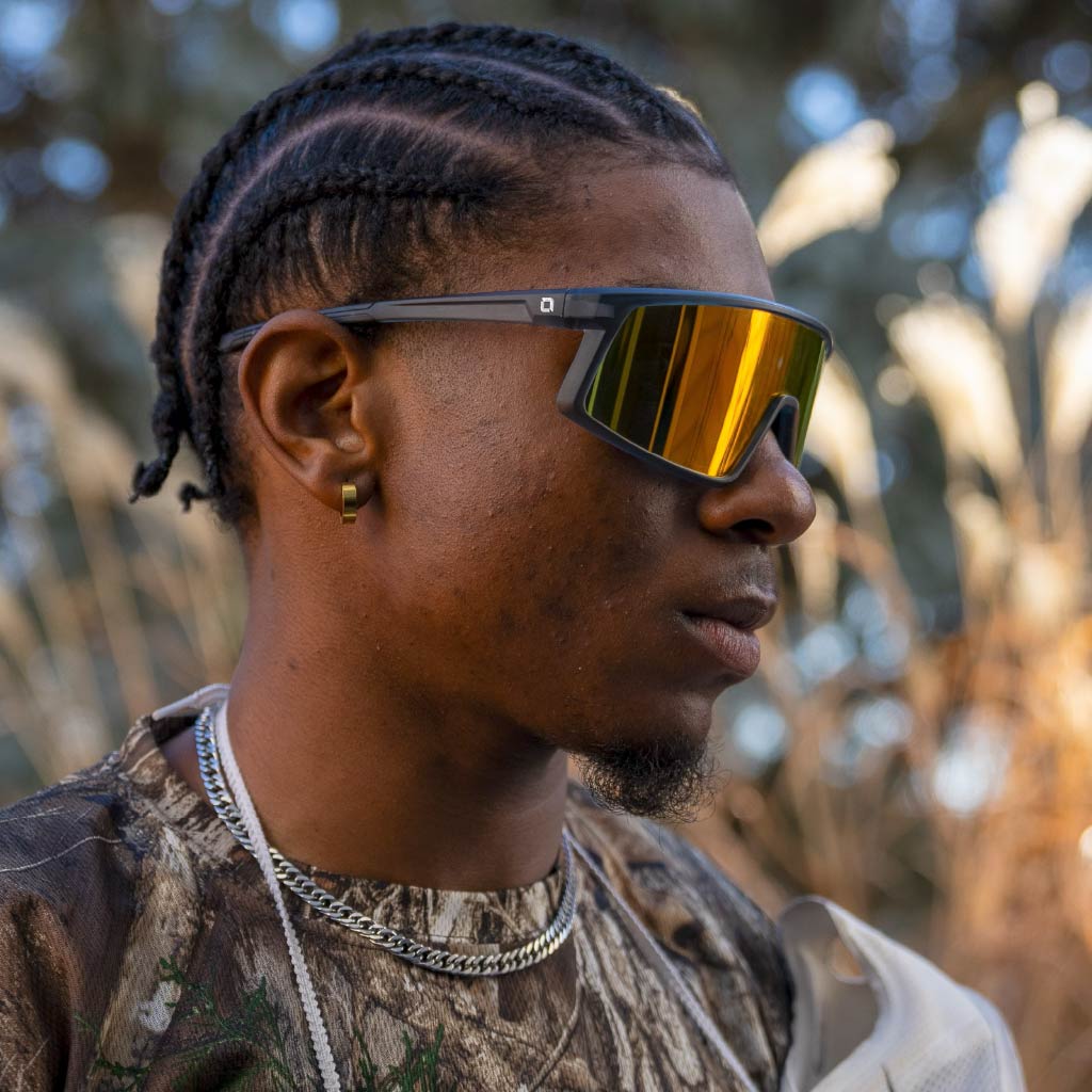 Person wearing Optic Nerve Stone Cat sunglasses with a gray frame, smoke lens, and red mirror, with a blurred natural background