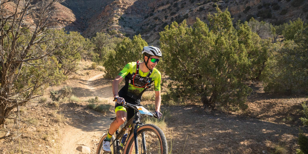 Person riding a mountain bike on a trail wearing Optic Nerve FixieMAX sunglasses with a gray frame, smoke lens, and green mirror, in a desert landscape 