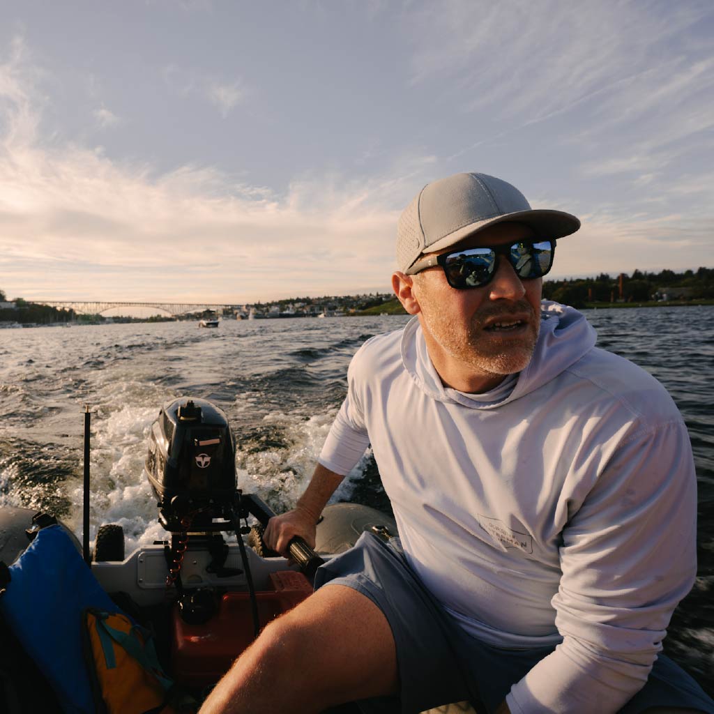 Man on a boat wearing Optic Nerve Zaldaingerous Rumble sunglasses with a black frame, amber lenses, and blue mirror, and a cap, with water and sky in the background