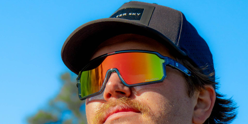 Man wearing a cap and Optic Nerve Scraggy sunglasses with a gray frame, smoke lens, and a red mirror, with a clear blue sky background