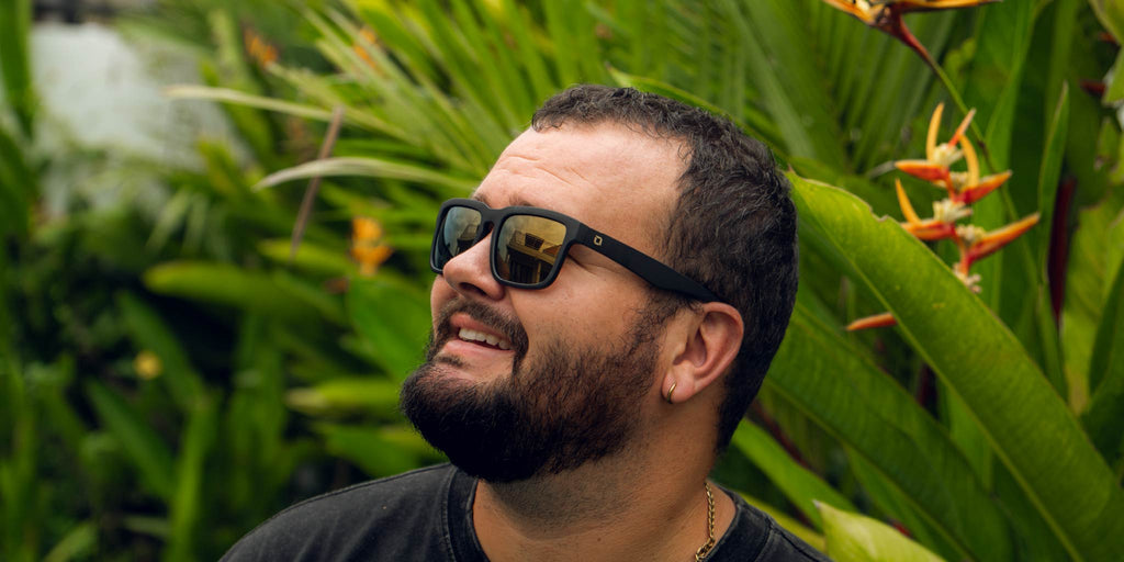 Man wearing Optic Nerve Mashup sunglasses with a black frame, brown lenses, and a gold mirror, with a tropical background