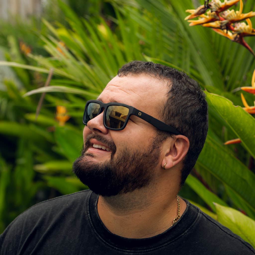Man wearing Optic Nerve Mashup sunglasses with a black frame, brown lenses, and a gold mirror, with a tropical background