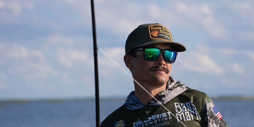 Man wearing a cap and Optic Nerve Ziggy sunglasses with a black frame, smoke lenses, and a green mirror, holding a fishing rod with a blurred background