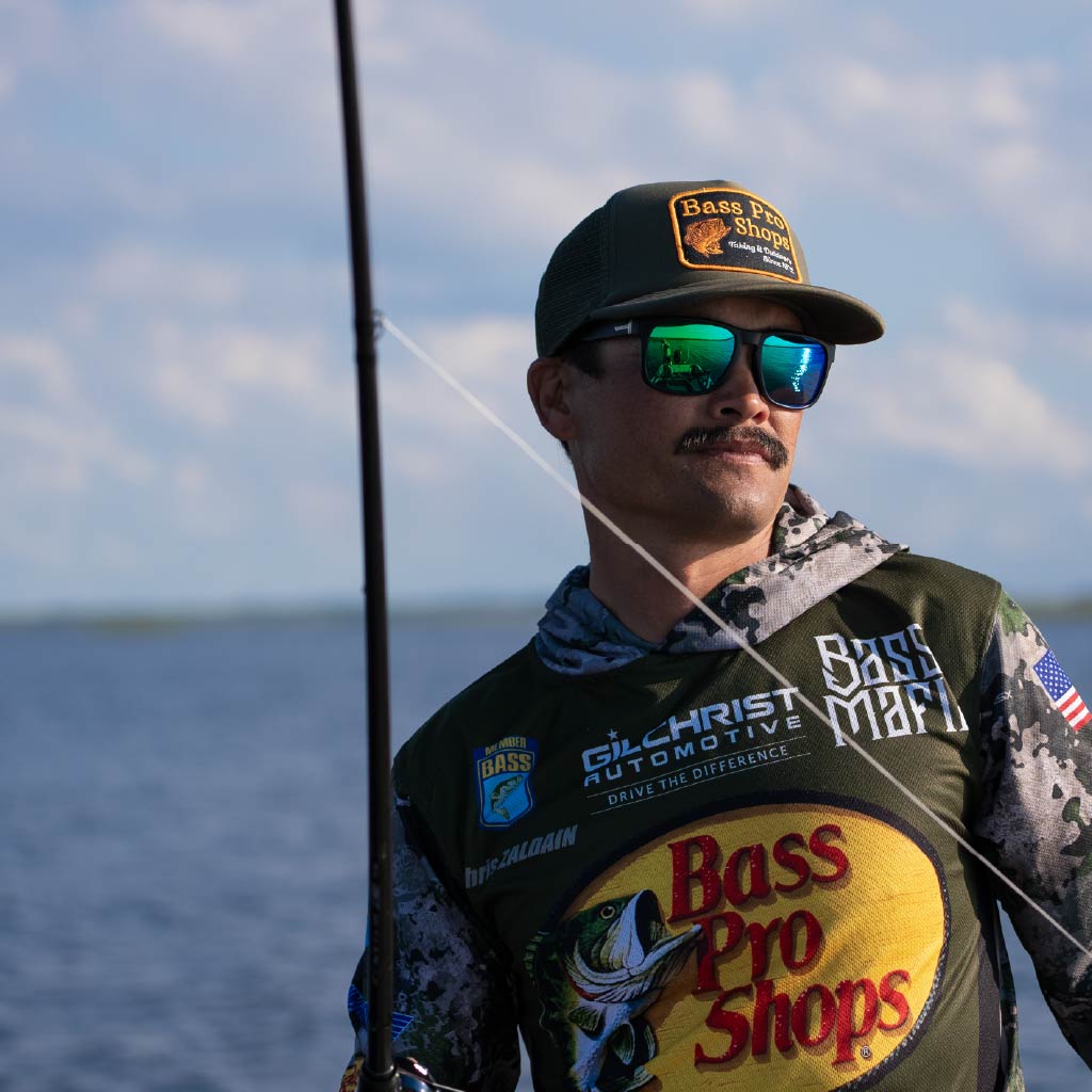 Man wearing a cap and Optic Nerve Ziggy sunglasses with a black frame, smoke lenses, and a green mirror, holding a fishing rod with a blurred background