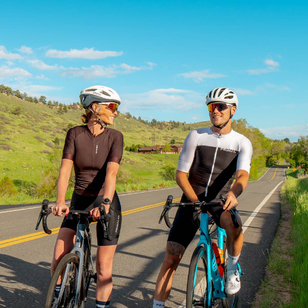 Two cyclists wearing Optic Nerve FixieAIR sunglasses with a white frame, smoke lens, and red mirror, on a road with a scenic background