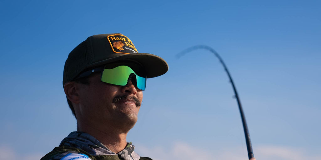 Man casting a fishing line wearing Optic Nerve Zaldaingerous FixieMAX sunglasses with a black frame, amber lens, and green mirror against a clear blue sky