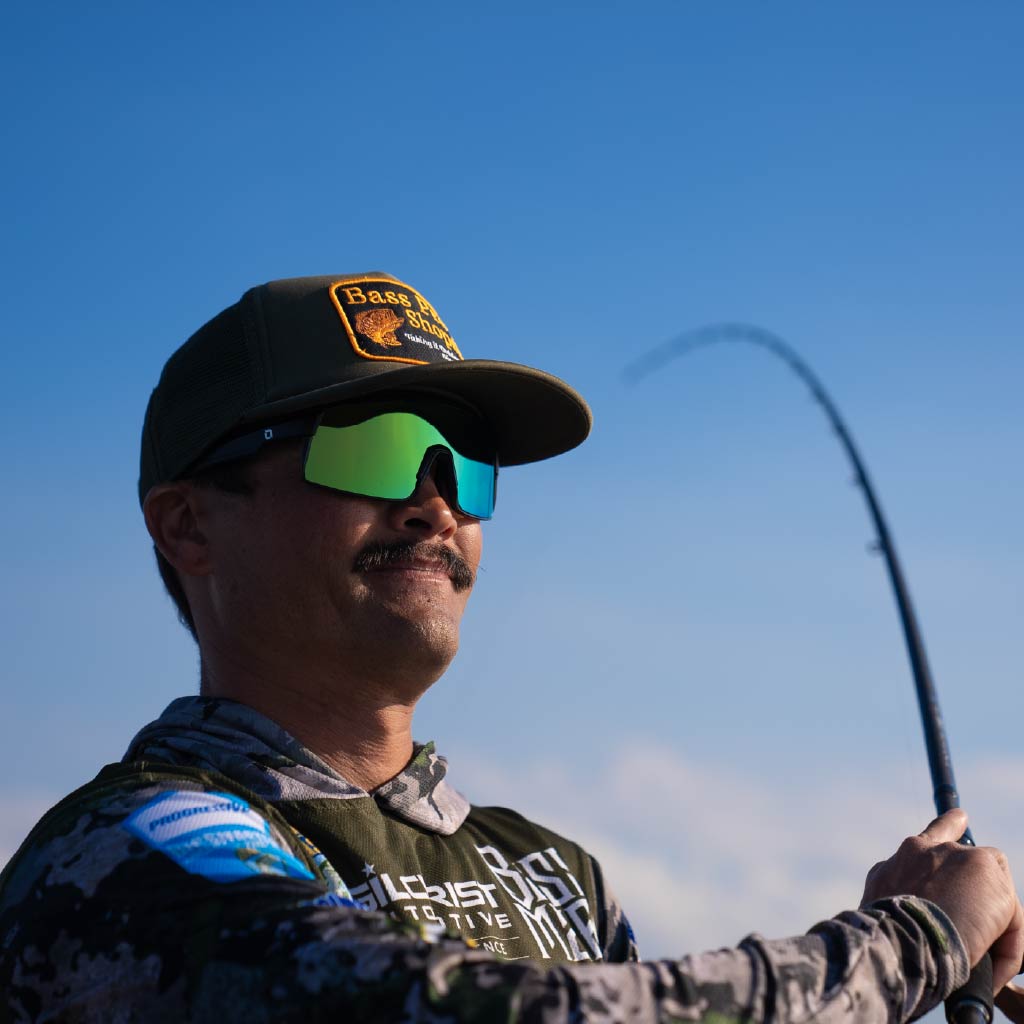 Man casting a fishing line wearing Optic Nerve Zaldaingerous FixieMAX sunglasses with a black frame, amber lens, and green mirror against a clear blue sky