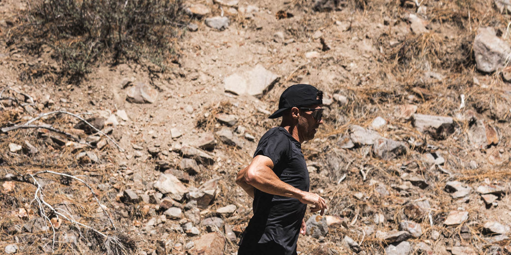 Person running on a rocky trail wearing Optic Nerve FixieAIR sunglasses with a black frame, smoke lens, and a silver mirror, in a desert-like environment
