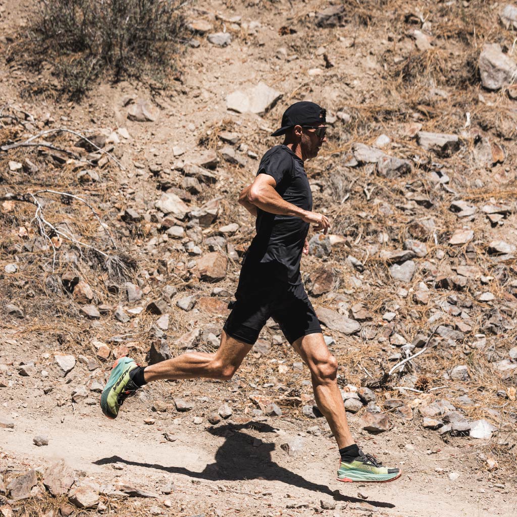 Person running on a rocky trail wearing Optic Nerve FixieAIR sunglasses with a black frame, smoke lens, and a silver mirror, in a desert-like environment