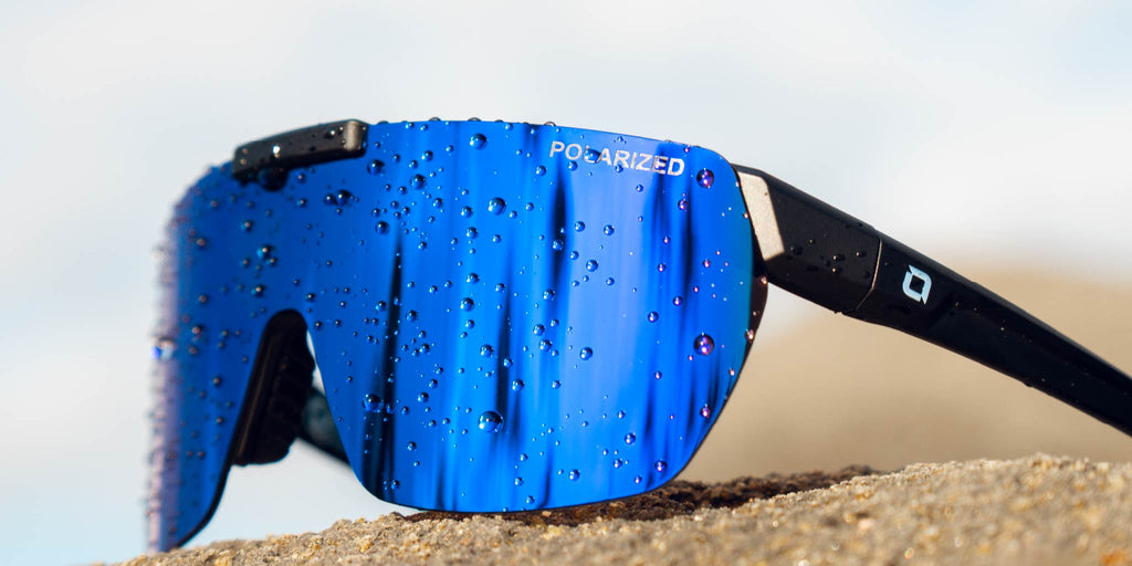 Close up of Optic Nerve Rockpile sunglasses with a black frame, smoke lens, and a blue mirror perched on a rock.