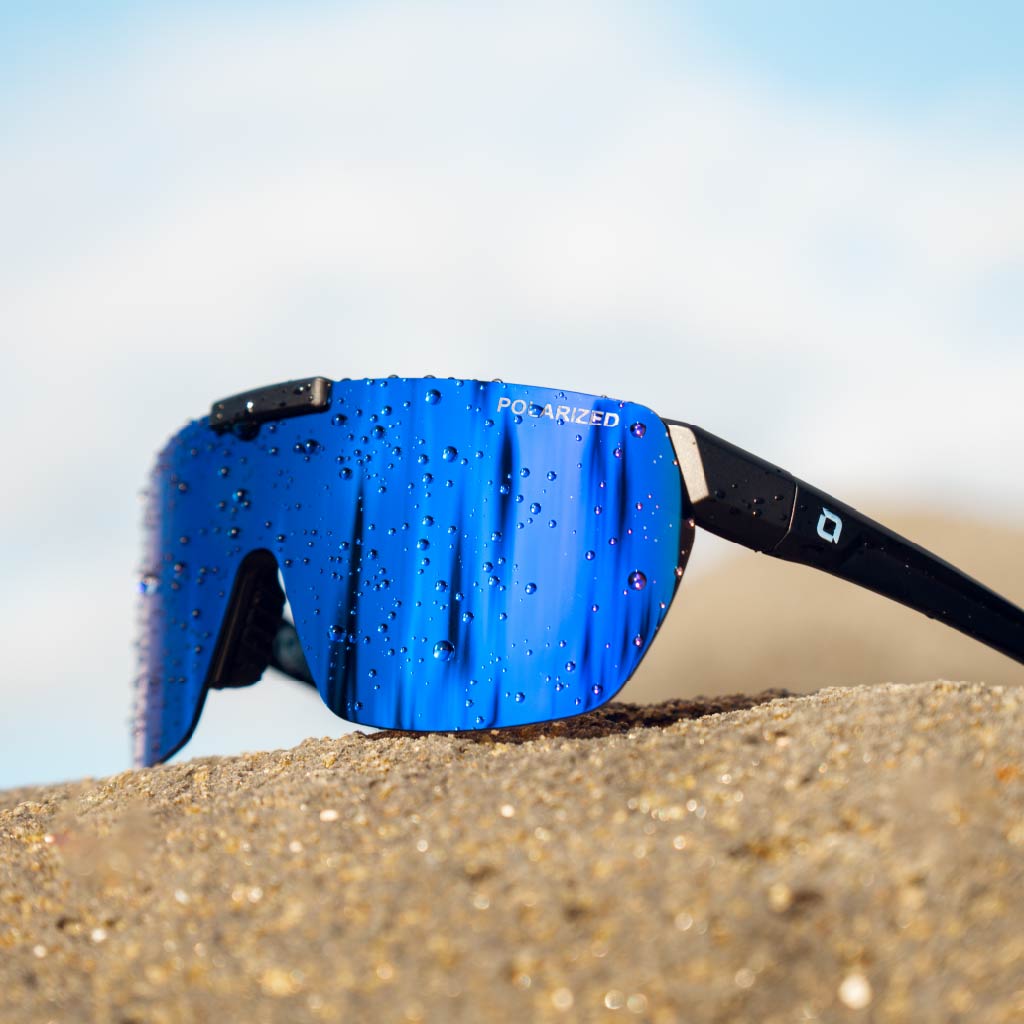 Close up of Optic Nerve Rockpile sunglasses with a black frame, smoke lens, and a blue mirror perched on a rock.