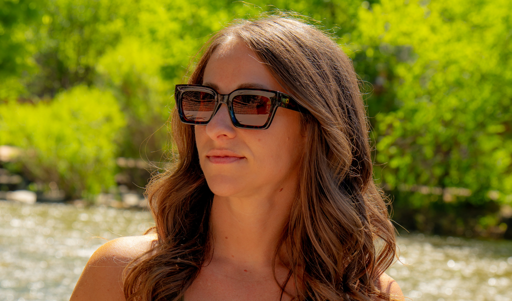 Woman wearing Optic Nerve Afterglow sunglasses with a tortoise frame and brown lenses, with a natural background of trees and water