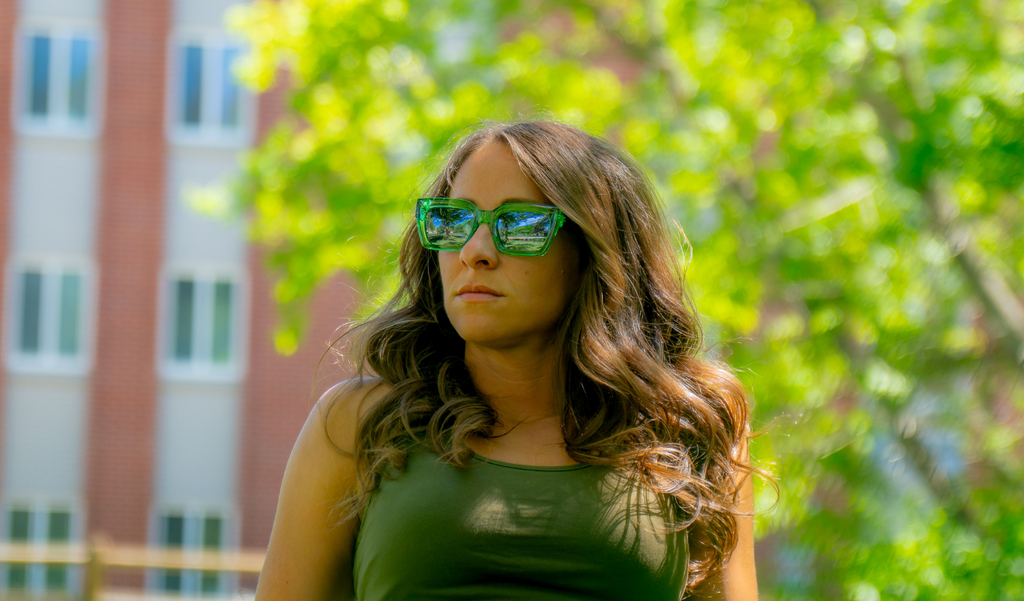 Woman wearing Optic Nerve Afterglow sunglasses with a green frame and smoke lenses, and a green top outdoors with trees and a building in the background