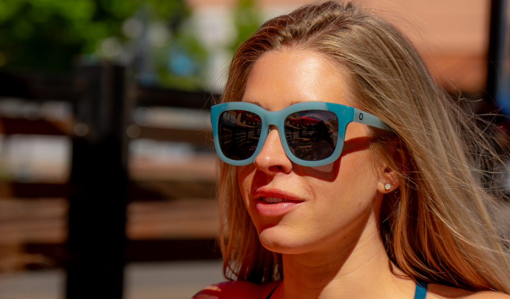 Woman wearing Optic Nerve Baby Doe sunglasses with a blue frame and smoke lenses with a blurred outdoor background