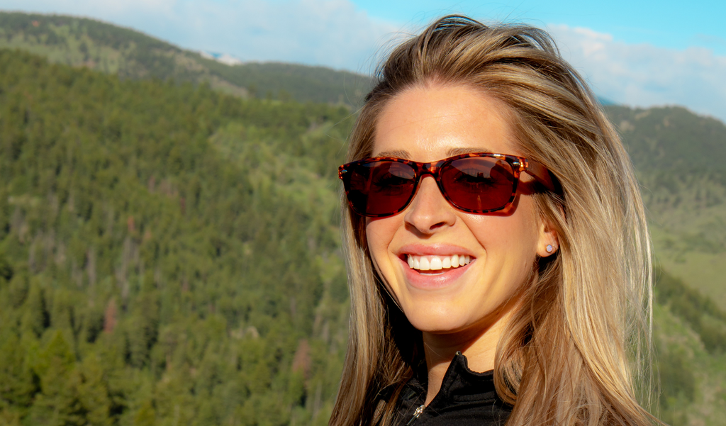 Woman wearing Optic Nerve Cruzin' Wood sunglasses with FSC natural wood temples, a tortoise frame, and brown lenses, with a scenic background of trees and mountains