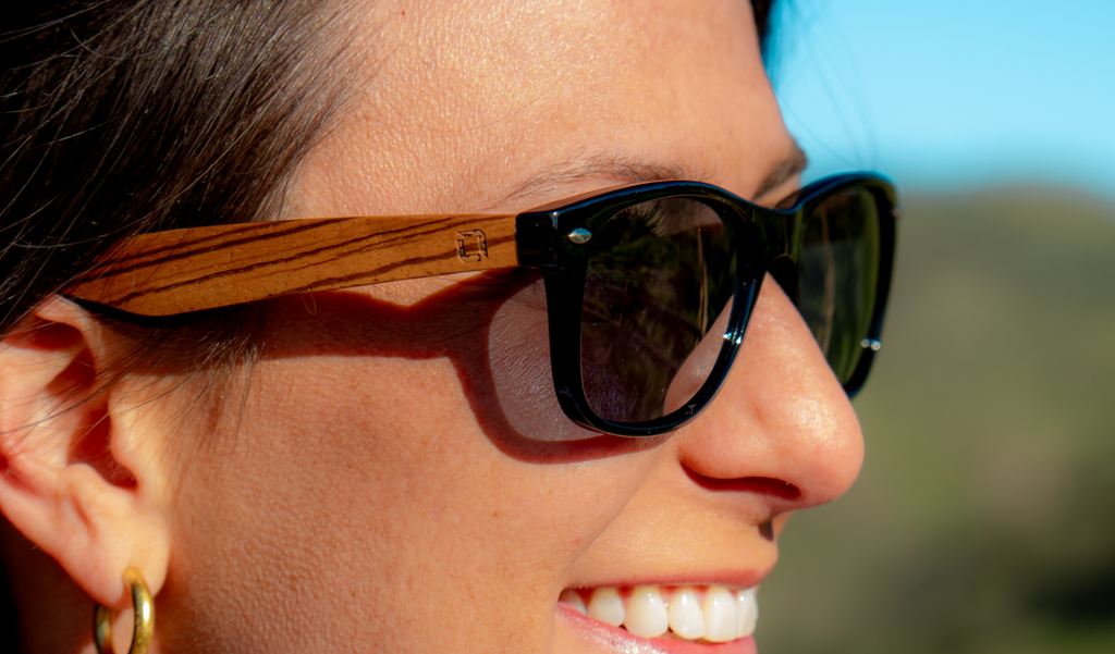 Close-up of a person wearing Optic Nerve Cruzin Wood sunglasses with FSC natural wood temples, black frame, and smoke lenses, with a blurred natural background