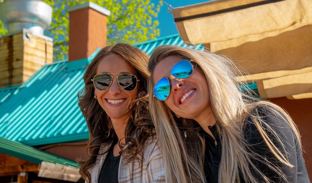 Two women wearing Optic Nerve Estrada sunglasses outdoors with a building and greenery in the background