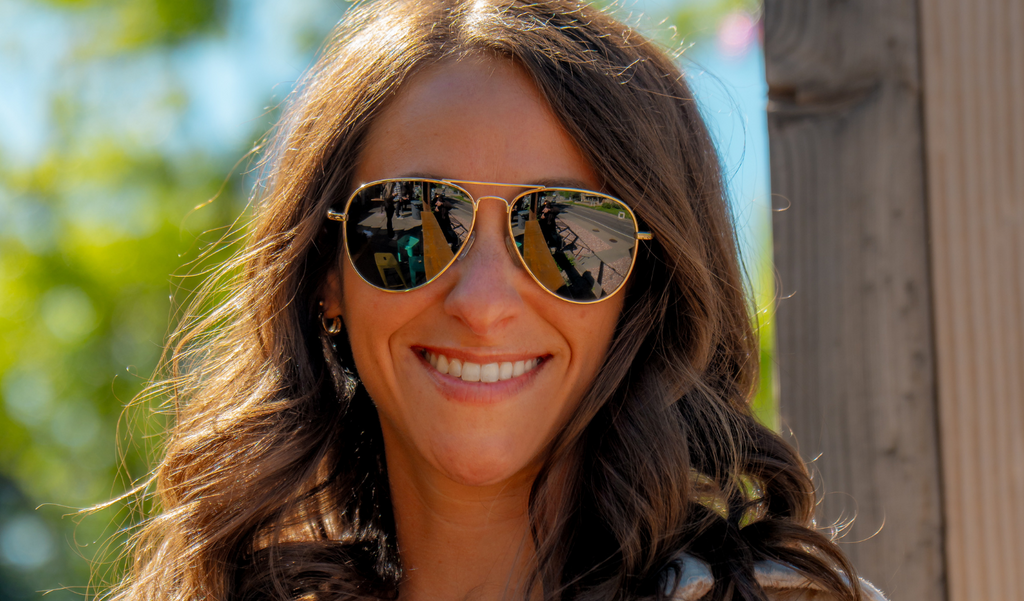 Woman wearing Optic Nerve Estrada  sunglasses with a gold frame and smoke lenses, with a blurred outdoor background