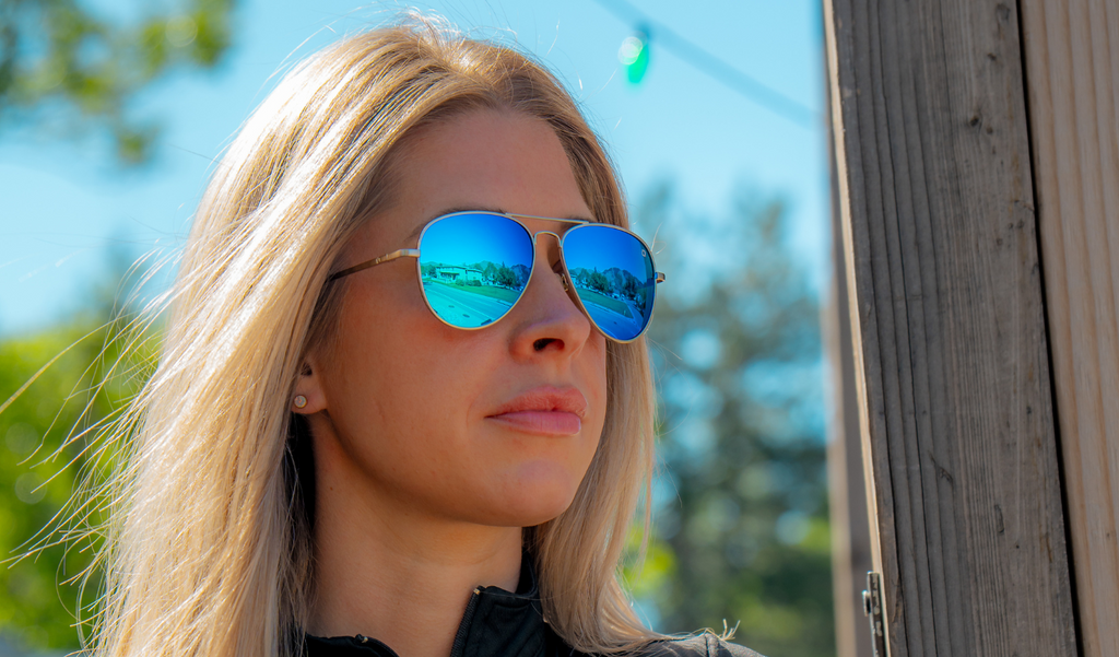 Woman wearing Optic Nerve Estrada sunglasses with a silver frame, brown lens, and blue mirror with a blurred outdoor background