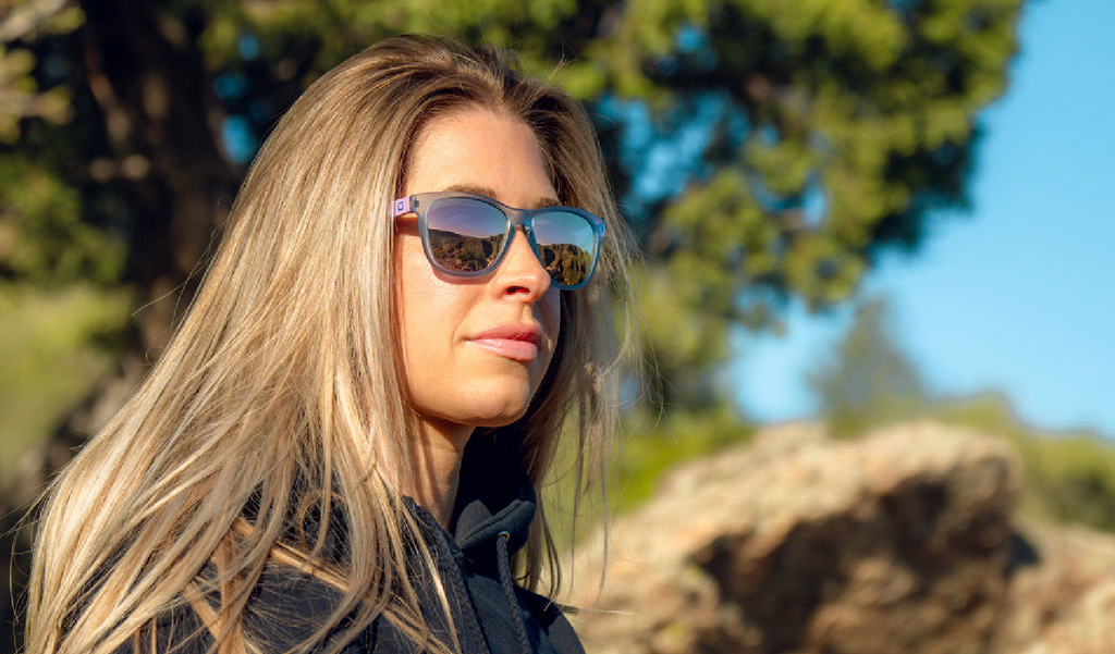 Woman wearing Optic nerve Fairplay sunglasses with a blue frame, smoke lenses, and a pink mirror, with a natural background of trees and rocks