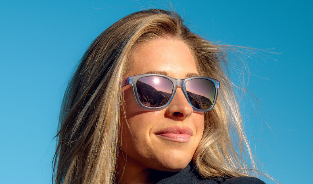 Woman wearing Optic Nerve Fairplay sunglasses with a blue frame, smoke lenses, and a pink mirror, with a clear blue sky background