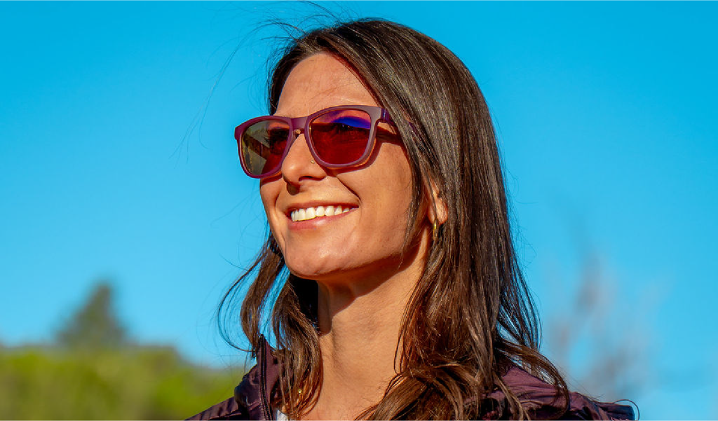 Woman wearing Optic Nerve Fairplay sunglasses with a purple frame, brown lenses, and a purple mirror, with a blue sky background