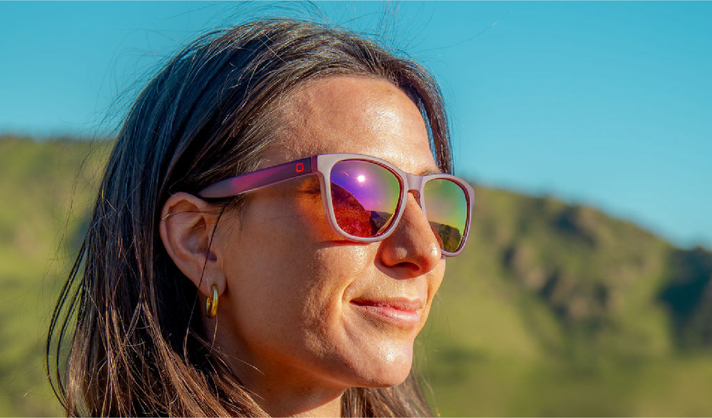 Woman wearing Optic Nerve Fairplay sunglasses with a purple frame, brown lenses, and a purple mirror, with a mountainous landscape in the background