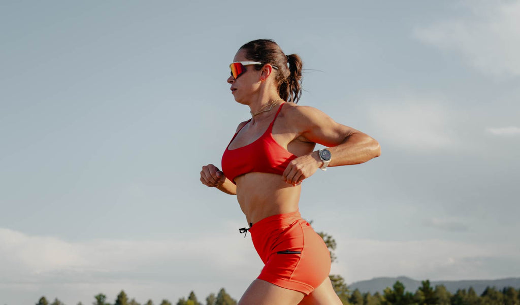 Woman in red athletic wear and Optic Nerve FixieAIR sunglasses with a white frame, smoke lens, and red mirror, running outdoors with a clear sky background