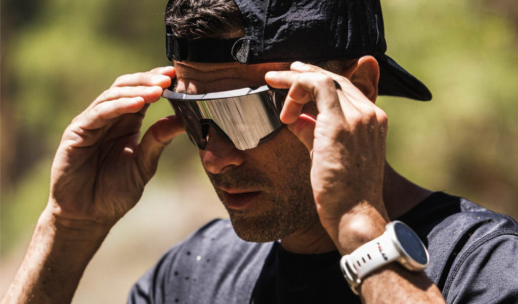 Person adjusting Optic Nerve FixieAIR sunglasses with a black frame, smoke lens, and silver mirror outdoors with a blurred natural background