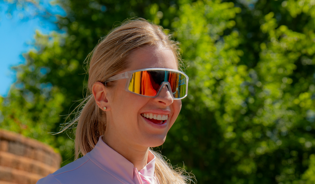 Person wearing Optic Nerve Gravel Cat sunglasses with a clear frame, smoke lens, and a red mirror with a blurred green outdoor background