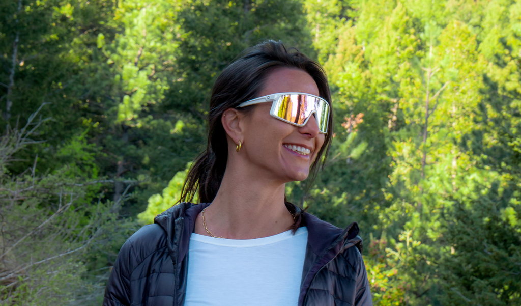 Woman wearing Optic Nerve Gravel Cat sunglasses with a white frame, brown lens, and a pink mirror, with a forest background
