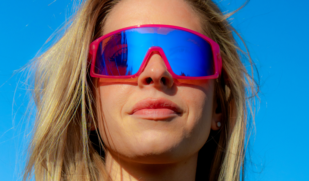 Person wearing Optic Nerve Gravel Cat sunglasses with a pink frame, brown lens, and blue mirror, against a clear blue sky