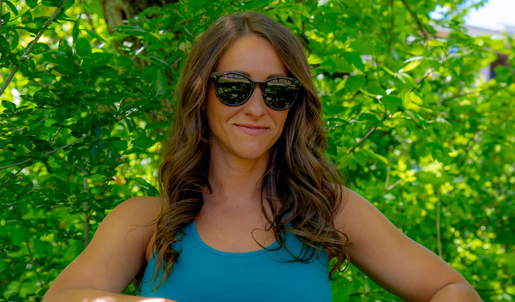 Woman wearing Optic Nerve Hotplate sunglasses with a tortoise frame and brown lenses, and a blue top in front of green foliage