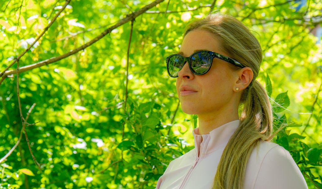 Woman wearing Optic Nerve Hotplate sunglasses with a black frame and smoke lenses, with a blurred green foliage background