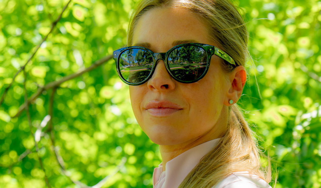 Woman wearing Optic Nerve Hotplate sunglasses with a black frame and smoke lenses, with a blurred green foliage background