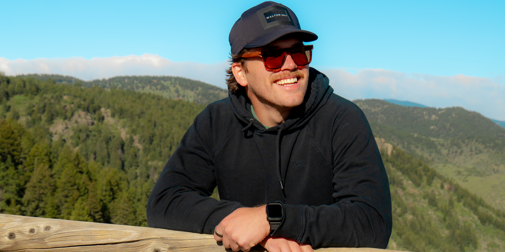 Person wearing a black jacket and Optic Nerve Mashup Wood sunglasses with FSC Natural Wood temples, tortoise frame, and brown lenses, with a scenic mountain background